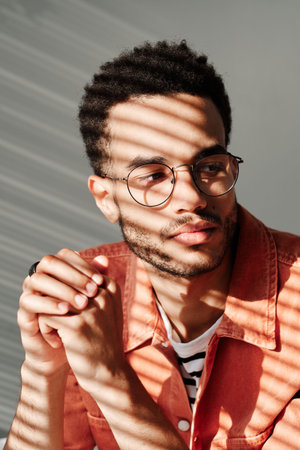 Pensive young black man with stubble wearing round glasses looking away, light through blinds effect