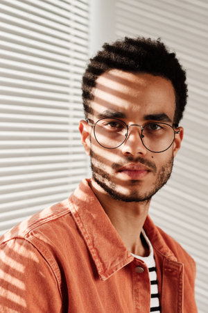 Portrait of serious young black man in round-shaped eyeglasses and coral shirt sitting against...