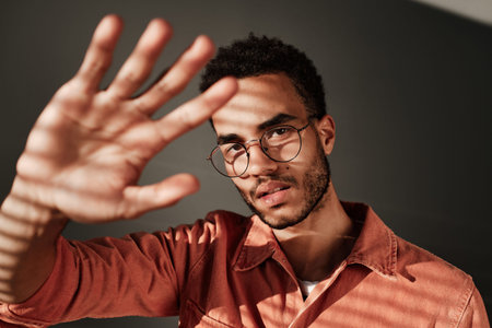 Portrait of serious handsome young black man in round-shaped glasses covering face from light with hand in dark roomの写真素材