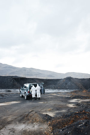Rear view of environmental scientists in white protective suit unloading van trunk on polluted land