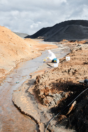 Pollution researcher in protective workwear crouching at river and taking sample of water while...