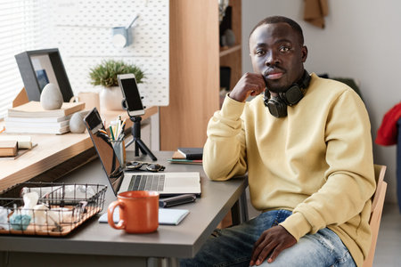 Portrait of serious young African American student sitting at desk wuth smartphone on tripod, laptop...