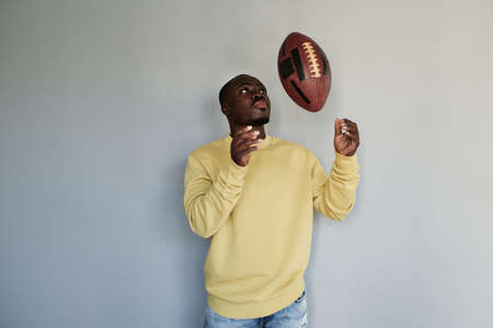 Serious young black rugby player of college team standing against gray wall and throwing ballの写真素材
