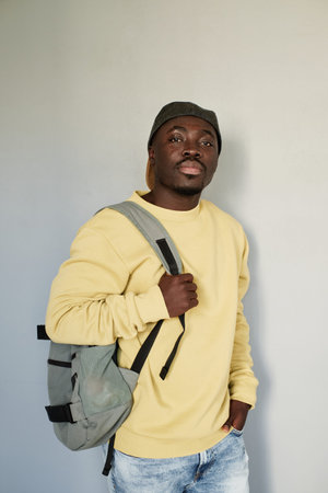 Portrait of serious handsome Black student guy in yellow sweatshirt and cap standing with satchel...