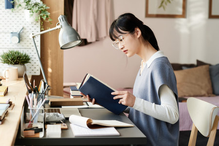 Concentrated Asian student girl in eyeglasses and knitted vest sitting at table and reading textbook...