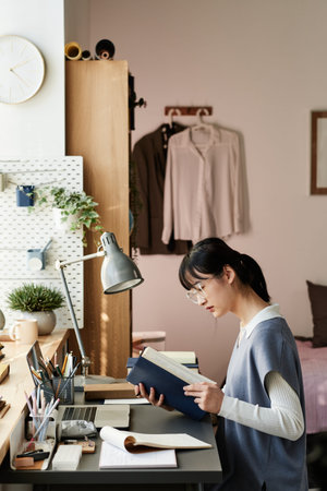 Serious curious Asian student girl in eyeglasses sitting at desk and reading textbook in room