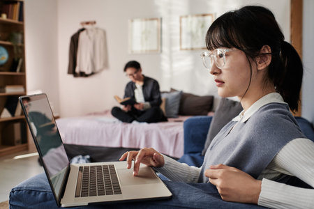 Concentrated Asian student girl in eyeglasses searching for information on laptop while her roommate...