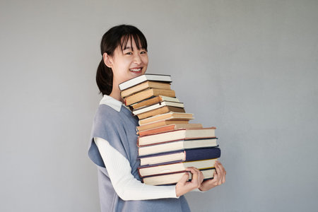 Portrait of positive Asian student girl standing with stack of books against gray background
