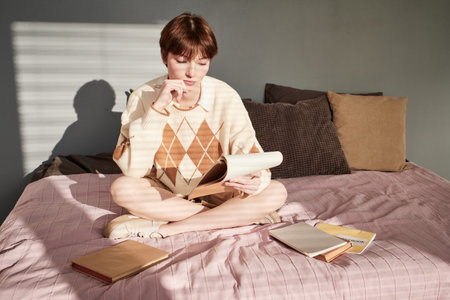 Thoughtful redhead student girl in sweater sitting on bed and reading notes while preparing for examの写真素材