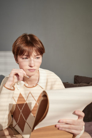 Serious redhead Caucasian student girl in knitted sweater holding pencil and analyzing notes in...