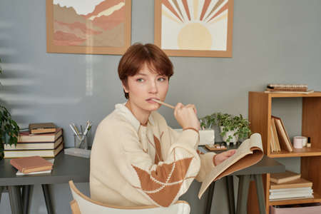 Serious thoughtful redhead Caucasian student girl with short hair keeping pencil in mouth while thinking about notes in sketchpadの写真素材