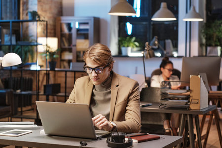 Young Caucasian man in eyeglasses and jacket sitting at desk and using laptop while working In Dark Officeの写真素材