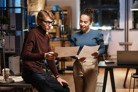 Content young multiethnic colleagues analyzing charts while working with papers in office at night