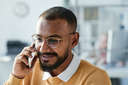Smiling handsome young mixed race business manager with beard talking to client by phone while...