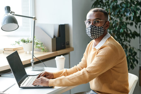 Portrait of serious young mixed race manager in checkered mask and eyeglasses sitting at table with...