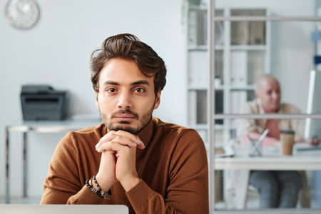 Portrait of serious handsome young middle eastern office manager in brown turtleneck sitting with clasped hands in officeの写真素材