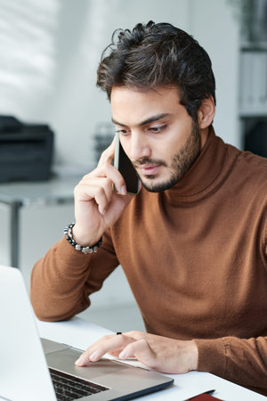 Serious modern businessman in brown turtleneck sitting at table and using laptop while having phone...