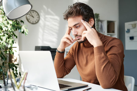 Serious young middle eastern man in brown turtleneck focused on thoughts looking at laptop screen...