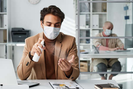 Young middle eastern employee in facial mask sitting at office table and applying spray disinfectant on handsの写真素材
