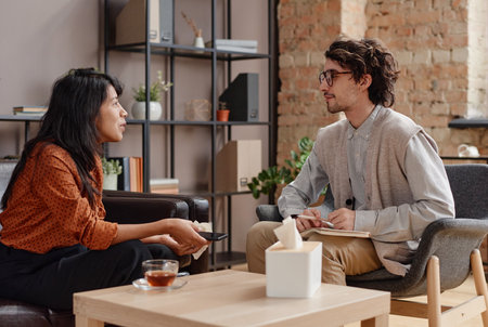 Horizontal medium shot of young adult woman sitting in front of professional psychologist telling him about successful momentsの写真素材