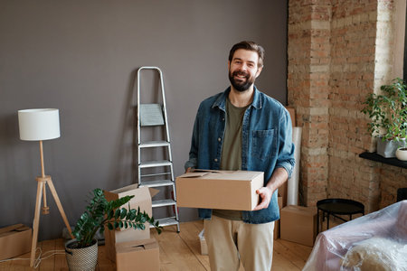 Horizontal shot of cheerful young man with beard on face wearing casual clothes holding box standing...