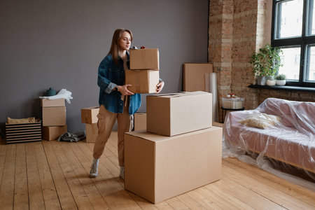 Horizontal shot of young woman moving to new apartment bringing and placing boxes with stuff in...
