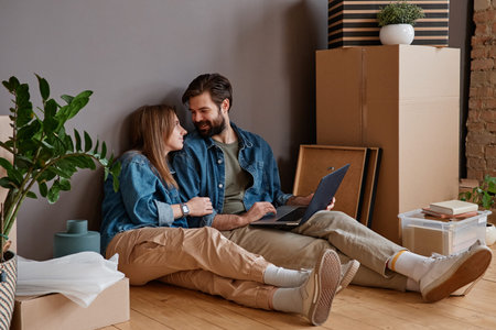 Young man and woman in love sitting on floor in new apartment surfing Internet on laptop searching...