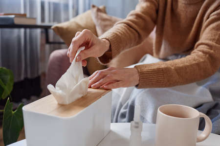 Horizontal close-up shot of unrecognizable woman having cold wearing warm clothes taking fresh paper napkin to blow noseの写真素材