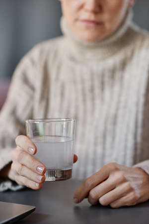 Vertical shot of mature woman having cold wearing sweater sitting at desk at home holding glass with mixture of water and effervescent tabletの写真素材