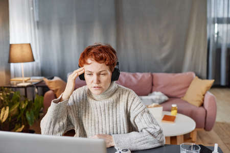 Mature woman sitting at desk in living room at home telling physician about headache during online consultationの写真素材