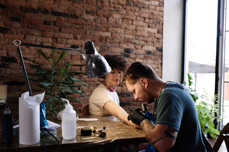 Professional male tattooist making hand tattoo with electric tattoo gun for young curly woman...