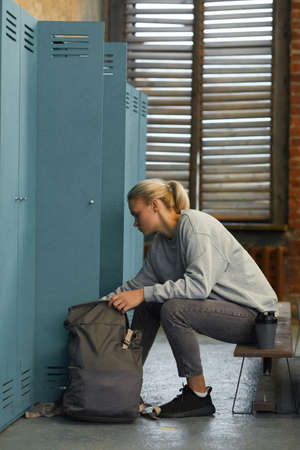 Vertical side view shot young woman sitting on bench in gym changeroom looking for something in her backpackの写真素材