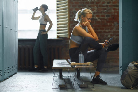 Two relaxed young women spending break time in gym dressing room horizontal shot