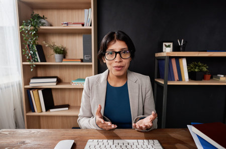 Portrait of confident Latin teacher in eyeglasses sitting in own office and gesturing hands while...