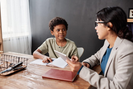 Young teacher using digital device while asking elementary student about topic at tutorial