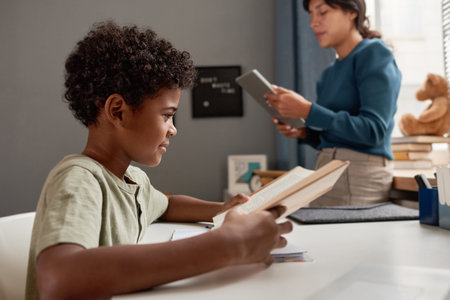 Content curious African American boy sitting at table and reading textbook while teacher using...