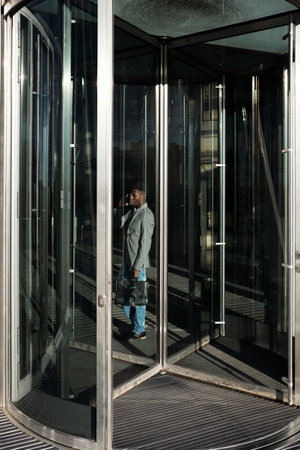 African American businessman standing behind glass revolving doors in business center and talking on...