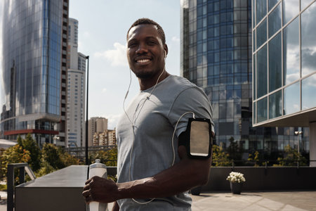 Waist-up portrait of handsome athletic Black man with earphones and smartphone in armband standing...
