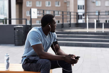 Young athletic African American man in sportswear resting on bench in city center and listening to music through earphones after workoutの写真素材