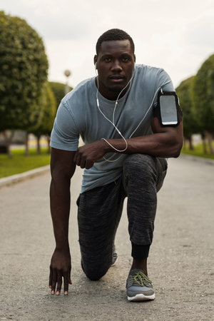 Runner getting ready to start. Portrait of young muscular Black sportsman with earphones standing on...