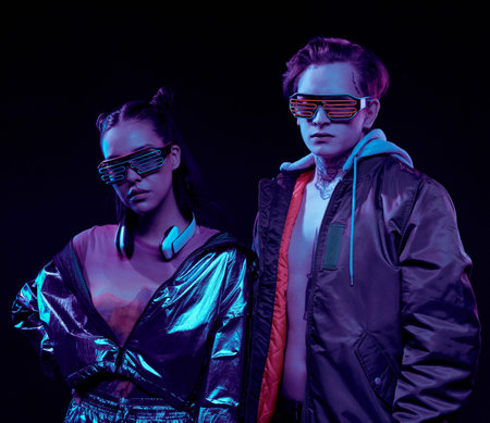 Two young people of cyber reality wearing stylish jackets and neon goggles standing against dark...