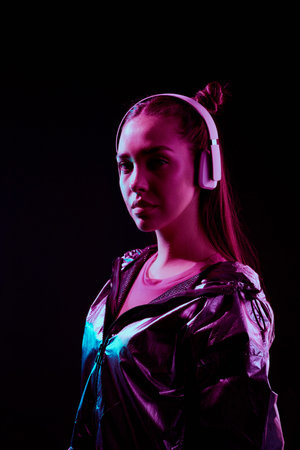 Portrait of serious stylish girl listening to music in wireless headphones while standing in...