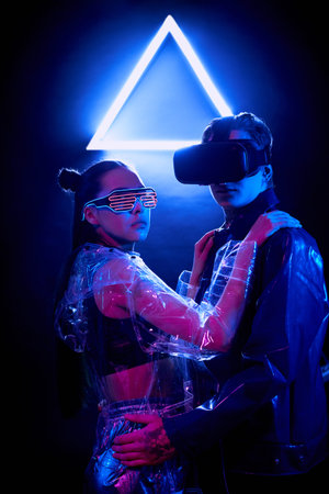 Portrait of serious young couple in virtual reality goggles embracing against neon triangle sign