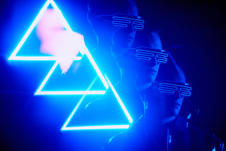 Cool boy in modern fashionable wire goggles standing in light of neon triangle signの写真素材