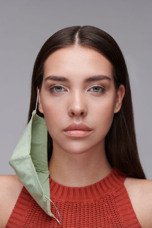 Portrait of serious young woman with cloth mask hanging on ear against gray background, coronavirus...