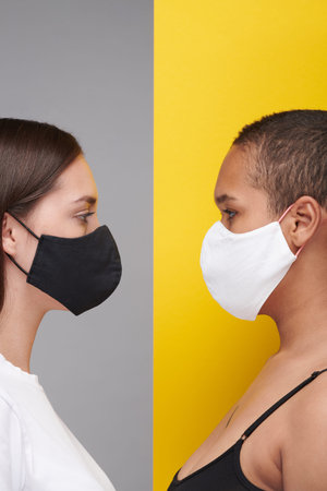 Young interracial women in black and white cloth masks looking at each other against gray an yellow background during coronavirusの写真素材