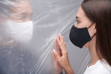 Young multi-ethnic women in cloth masks touching through polyethylene wall while having barrier during coronavirus epidemicの写真素材
