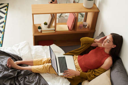 Serious exhausted young woman with closed eyes suffering from migraine and lying with laptop on bedの写真素材