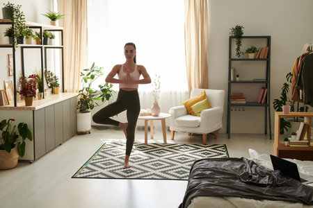 Serene young woman in sportswear standing on one leg while practicing tree pose in cozy living room...
