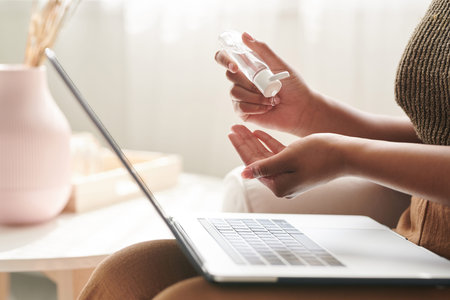 Close-up of unrecognizable woman sitting with laptop on knees and pouring antiseptic in hand while...